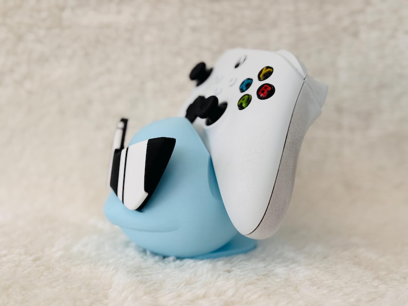 Puede incluir: Un soporte de mando azul claro con un mando Xbox blanco. El soporte tiene una forma redondeada con detalles en blanco y negro. El mando es blanco con botones de colores.