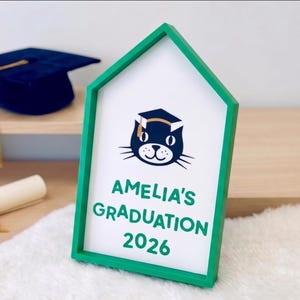 May include: A green framed sign with the text "AMELIA'S GRADUATION 2026" and a cat wearing a graduation cap. A rolled diploma with a gold ribbon and a navy blue graduation cap with a gold tassel are also visible.