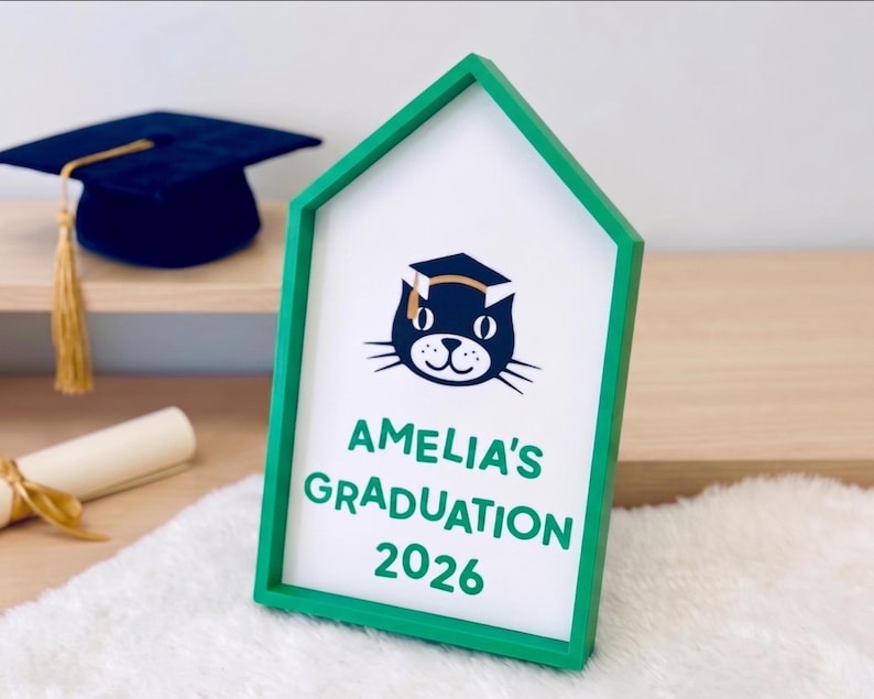 May include: A green framed sign with the text "AMELIA'S GRADUATION 2026" and a cat wearing a graduation cap. A rolled diploma with a gold ribbon and a navy blue graduation cap with a gold tassel are also visible.