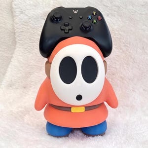 May include: A black video game controller resting on top of a Shy Guy character figure. The figure has a white oval mask with black eye and mouth holes, a coral-orange hooded robe, a brown belt with a yellow buckle, and solid blue rounded feet.