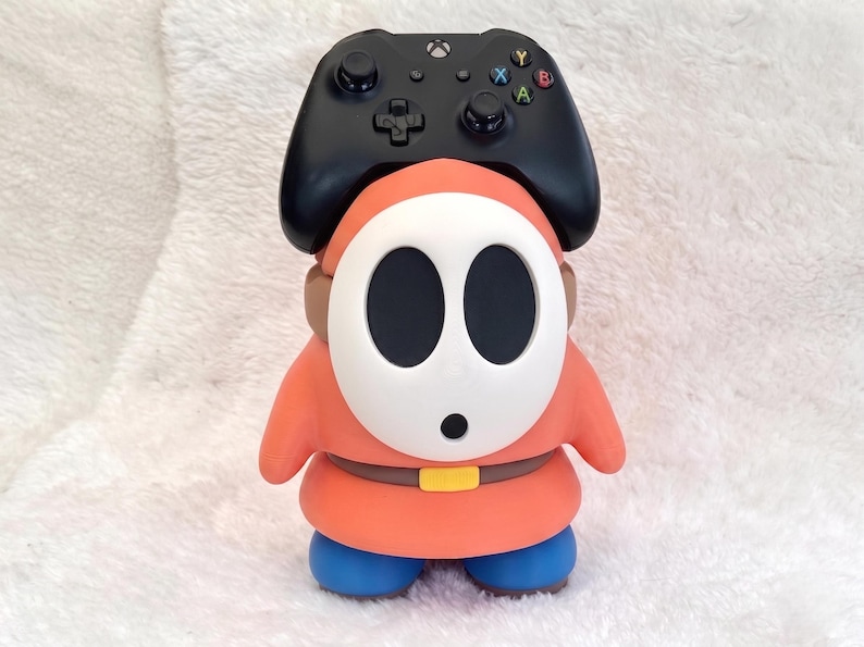May include: A black video game controller resting on top of a Shy Guy character figure. The figure has a white oval mask with black eye and mouth holes, a coral-orange hooded robe, a brown belt with a yellow buckle, and solid blue rounded feet.