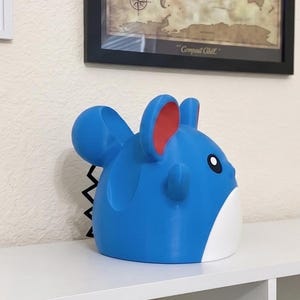 May include: A blue and white figurine of a cartoon character with large ears and a black zig-zag tail. The figurine has red accents on the ears and a black eye. The figurine is sitting on a white shelf.
