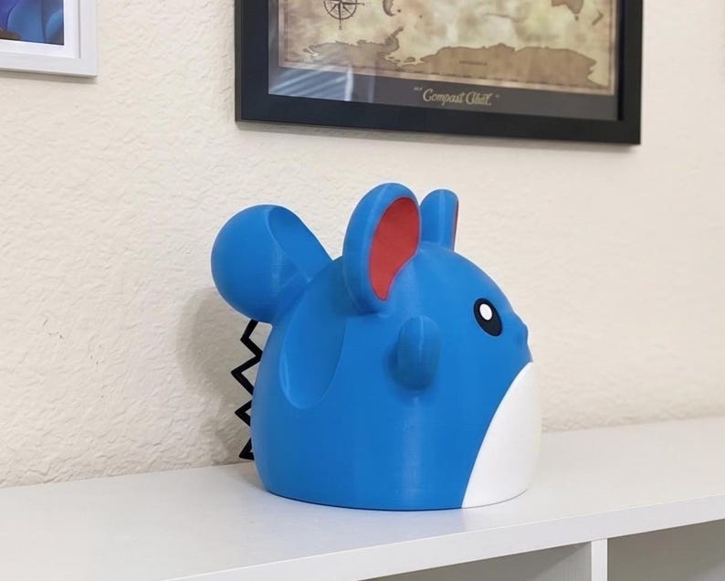 May include: A blue and white figurine of a cartoon character with large ears and a black zig-zag tail. The figurine has red accents on the ears and a black eye. The figurine is sitting on a white shelf.