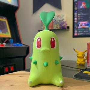 May include: A green Chikorita figure with red eyes and a green leaf on its head. The figure has small green spikes and is sitting on a wooden surface. A yellow Pikachu figure is in the background.
