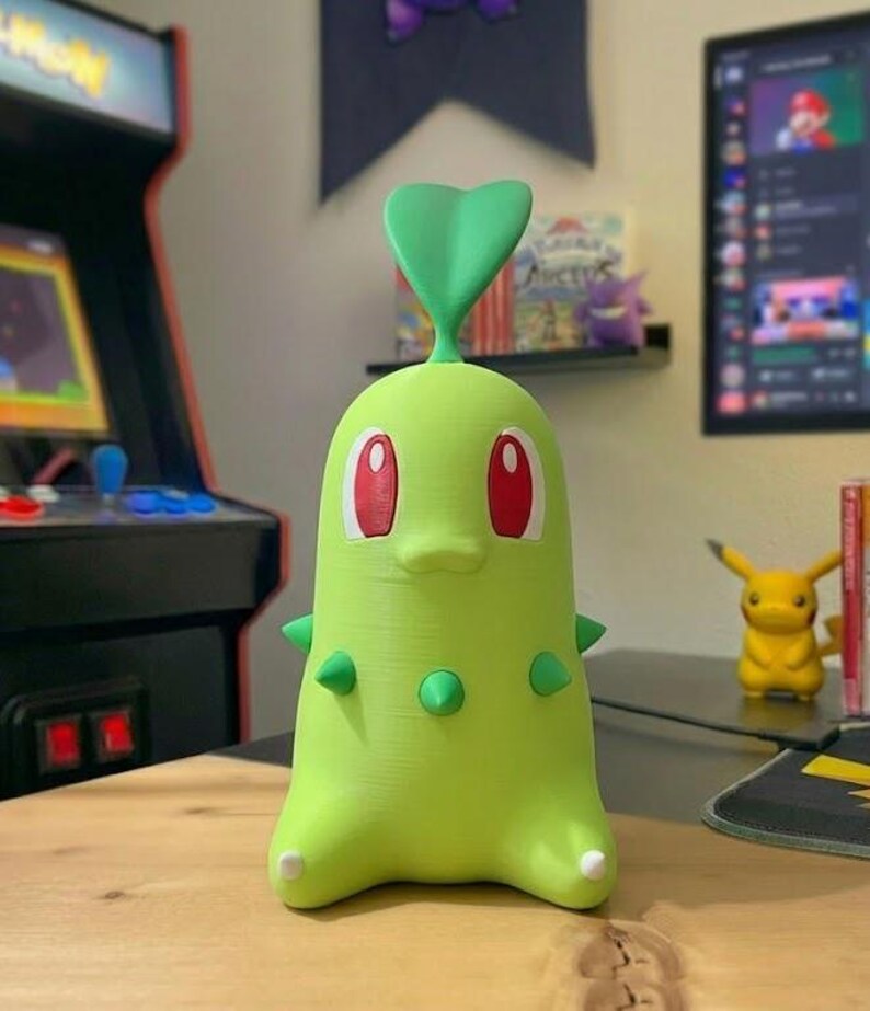 May include: A green Chikorita figure with red eyes and a green leaf on its head. The figure has small green spikes and is sitting on a wooden surface. A yellow Pikachu figure is in the background.