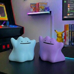 Pode incluir: Duas figuras em forma de Ditto, uma azul clara e outra roxa, est&atilde;o lado a lado. Cada uma tem um rosto sorridente simples. O fundo inclui um jogo de arcade retr&ocirc; e uma figura Pikachu.