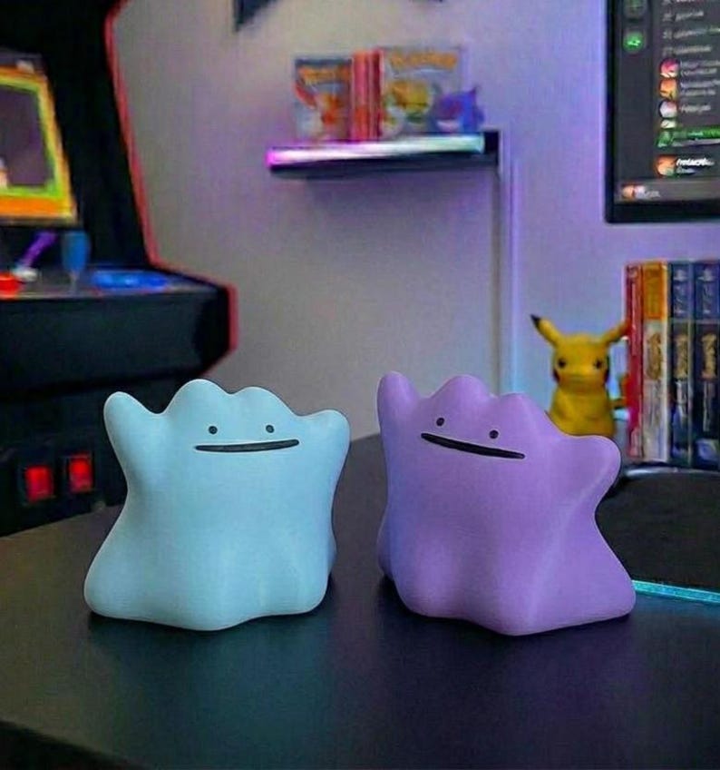 Pode incluir: Duas figuras em forma de Ditto, uma azul clara e outra roxa, est&atilde;o lado a lado. Cada uma tem um rosto sorridente simples. O fundo inclui um jogo de arcade retr&ocirc; e uma figura Pikachu.