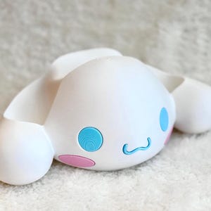 Puede incluir: Un soporte para tel&eacute;fono de pl&aacute;stico blanco con forma de personaje, que presenta grandes ojos azules circulares, una peque&ntilde;a boca azul curvada y acentos ovalados de color rosa en las mejillas. Incluye dos extensiones laterales redondeadas.