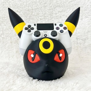 May include: A black and yellow controller stand shaped like the head of the creature Umbreon, featuring pointed ears, red eyes, and a yellow forehead ring. A white game controller is resting securely on top of the character-themed display stand.