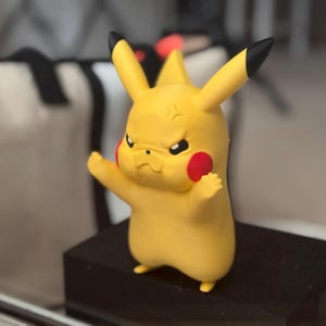 May include: A yellow Pikachu figurine with a frowning expression, black-tipped ears, and red circular cheeks. The character is depicted in a standing pose with arms slightly raised, featuring a smooth matte finish and distinct facial features.