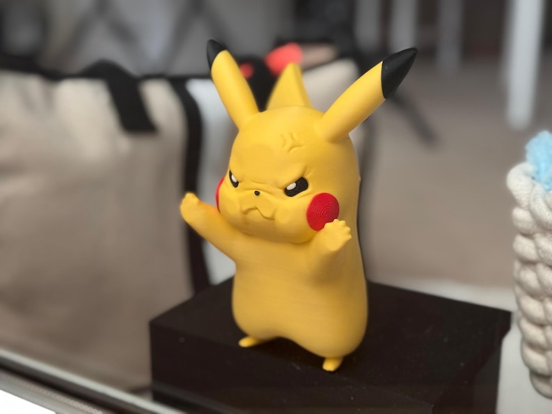 May include: A yellow Pikachu figurine with a frowning expression, black-tipped ears, and red circular cheeks. The character is depicted in a standing pose with arms slightly raised, featuring a smooth matte finish and distinct facial features.