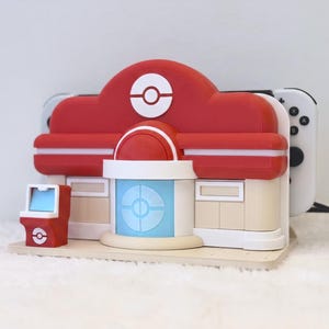 May include: A red and beige plastic charging dock shaped like a Pokemon Center building, featuring a central blue door, a small red arcade cabinet on the left, and a white Poke Ball emblem on the roof, holding a white gaming console.
