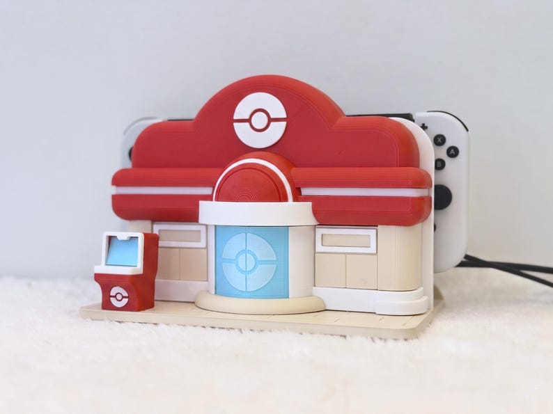 May include: A red and beige plastic charging dock shaped like a Pokemon Center building, featuring a central blue door, a small red arcade cabinet on the left, and a white Poke Ball emblem on the roof, holding a white gaming console.