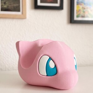 May include: A pink, 3D-printed figure of a cartoon character with large blue and white eyes. The figure has a rounded shape with a small ear on top. The object is sitting on a white surface.