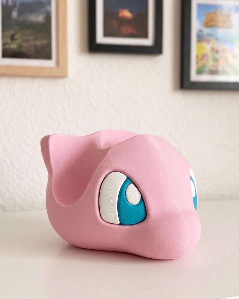 May include: A pink, 3D-printed figure of a cartoon character with large blue and white eyes. The figure has a rounded shape with a small ear on top. The object is sitting on a white surface.