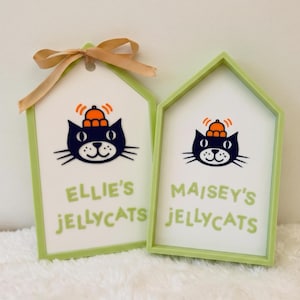 Puede incluir: Dos letreros en forma de casa con marcos verde claro y centros blancos. Cada uno presenta una ilustraci&oacute;n de un gato negro con un sombrero naranja y el texto "ELLIE'S JELLYCATS" o "MAISEY'S JELLYCATS" en verde claro. El letrero izquierdo tiene un lazo de cinta beige.
