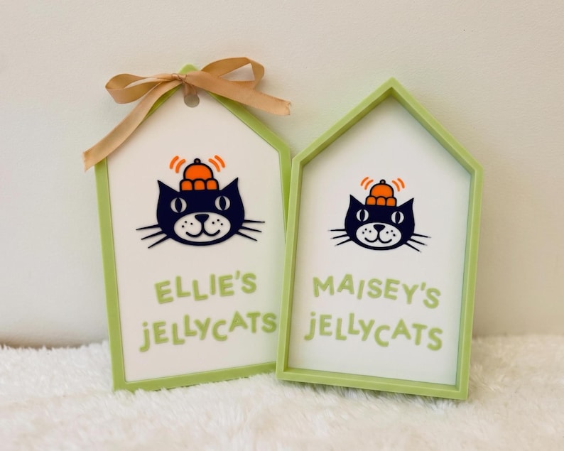 Puede incluir: Dos letreros en forma de casa con marcos verde claro y centros blancos. Cada uno presenta una ilustraci&oacute;n de un gato negro con un sombrero naranja y el texto "ELLIE'S JELLYCATS" o "MAISEY'S JELLYCATS" en verde claro. El letrero izquierdo tiene un lazo de cinta beige.