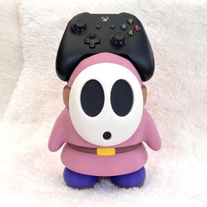 May include: A pink Shy Guy character figure serving as a controller stand, featuring a white oval mask with black eye holes, a brown belt with a yellow buckle, and blue shoes. A black Xbox wireless controller rests on top of the figure's head.