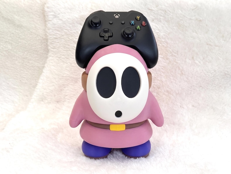 May include: A pink Shy Guy character figure serving as a controller stand, featuring a white oval mask with black eye holes, a brown belt with a yellow buckle, and blue shoes. A black Xbox wireless controller rests on top of the figure's head.