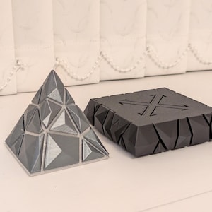 May include: A gray geometric pyramid puzzle and a black, angular device with an X-shaped design and arrows. The pyramid is composed of triangular and diamond-shaped pieces. The device has a power cord attached.
