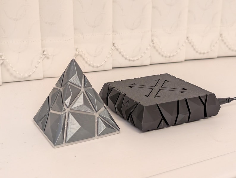 May include: A gray geometric pyramid puzzle and a black, angular device with an X-shaped design and arrows. The pyramid is composed of triangular and diamond-shaped pieces. The device has a power cord attached.