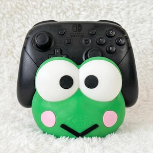 Puede incluir: Un mando Nintendo Switch Pro negro sobre un soporte con forma de rana verde. El soporte presenta grandes ojos blancos con pupilas negras, mejillas circulares rosas y una boca negra en forma de V para sostener el mando de juego de forma segura.