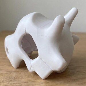 May include: A white, 3D-printed sculpture of a Cubone skull. The skull has a smooth, matte finish and features prominent eye sockets, a nasal cavity, and two horn-like protrusions. The sculpture is a unique piece of fan art.