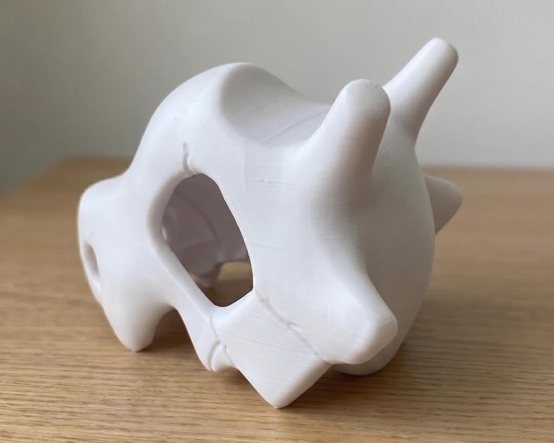 May include: A white, 3D-printed sculpture of a Cubone skull. The skull has a smooth, matte finish and features prominent eye sockets, a nasal cavity, and two horn-like protrusions. The sculpture is a unique piece of fan art.
