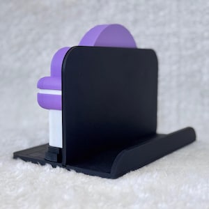 May include: A black plastic stand featuring a flat base and a vertical back panel. A purple and white object with a rounded top is positioned against the front of the black vertical panel, resting securely on the flat base of the stand.
