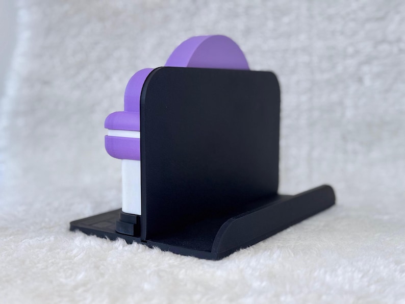 May include: A black plastic stand featuring a flat base and a vertical back panel. A purple and white object with a rounded top is positioned against the front of the black vertical panel, resting securely on the flat base of the stand.