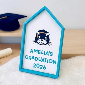 May include: A blue house-shaped frame with a white interior featuring a cat wearing a graduation cap. The text "AMELIA'S GRADUATION 2026" is displayed in blue. A graduation cap and diploma are visible in the background.