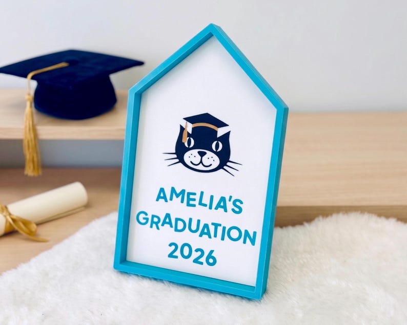 May include: A blue house-shaped frame with a white interior featuring a cat wearing a graduation cap. The text "AMELIA'S GRADUATION 2026" is displayed in blue. A graduation cap and diploma are visible in the background.
