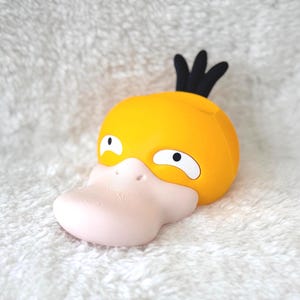 Puede incluir: Una cabeza de personaje amarilla similar a un pato con un pico rosa p&aacute;lido, ojos blancos con pupilas negras y tres mechones de pelo negro en la parte superior. El objeto tiene un acabado mate liso y una forma redondeada, parecida a una criatura estilizada.