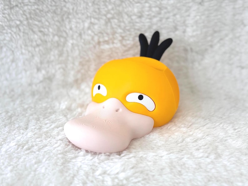 Puede incluir: Una cabeza de personaje amarilla similar a un pato con un pico rosa p&aacute;lido, ojos blancos con pupilas negras y tres mechones de pelo negro en la parte superior. El objeto tiene un acabado mate liso y una forma redondeada, parecida a una criatura estilizada.