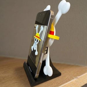 May include: A black smartphone stand featuring a white and yellow cartoon character design. The stand holds a smartphone displaying a skull and crossbones graphic. The stand is on a wooden surface.