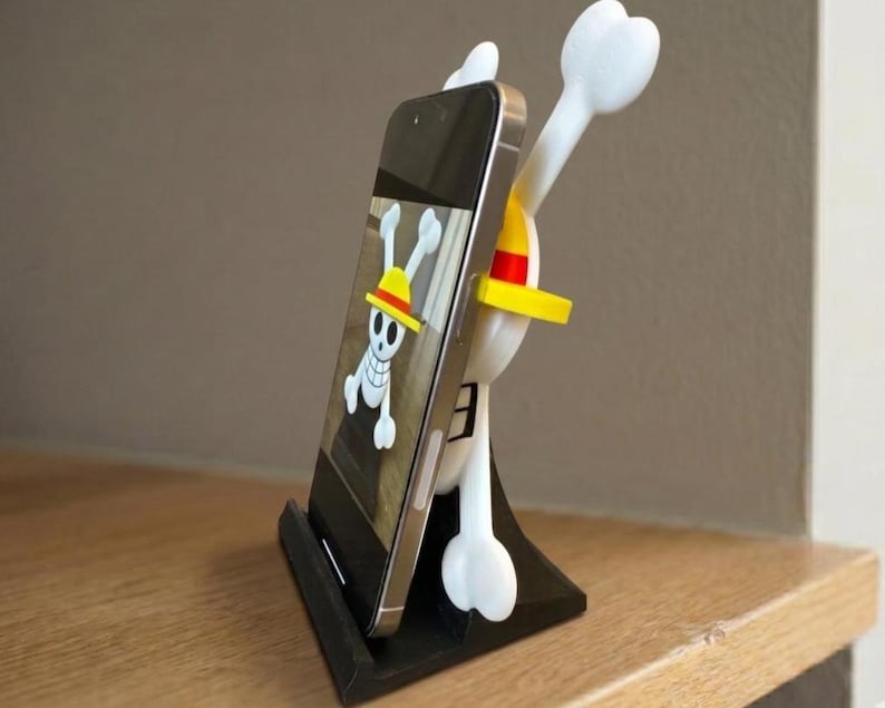 May include: A black smartphone stand featuring a white and yellow cartoon character design. The stand holds a smartphone displaying a skull and crossbones graphic. The stand is on a wooden surface.