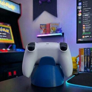 May include: A white PlayStation 5 controller on a blue charging stand. The controller features black joysticks and buttons. The background includes an arcade cabinet, a computer monitor, and a shelf with video games.