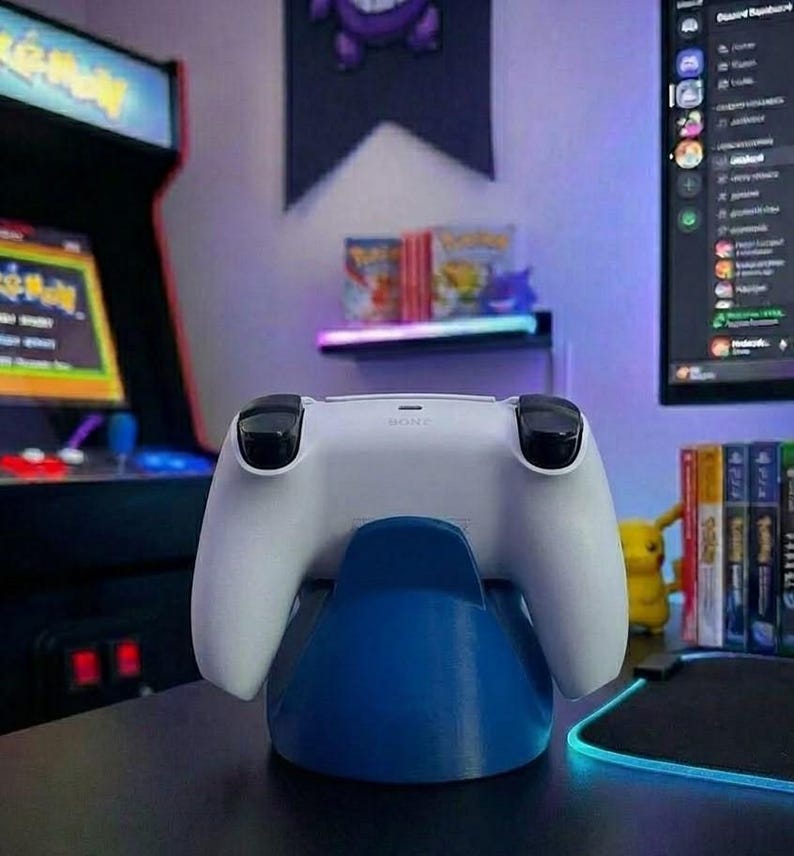 May include: A white PlayStation 5 controller on a blue charging stand. The controller features black joysticks and buttons. The background includes an arcade cabinet, a computer monitor, and a shelf with video games.