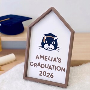 May include: A graduation-themed sign in a house-shaped frame. The sign features a cat wearing a graduation cap, the text "AMELIA'S GRADUATION 2026", a graduation cap, and a diploma. The sign is on a white fluffy surface.