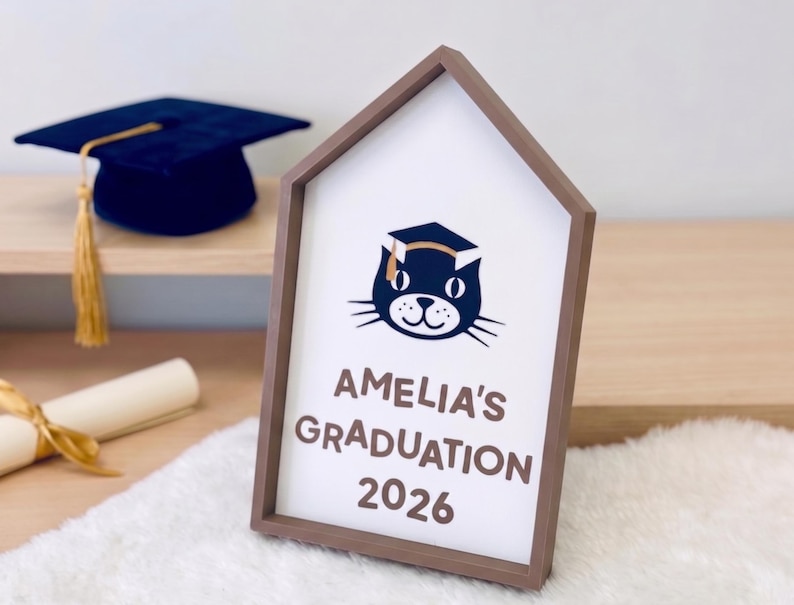 May include: A graduation-themed sign in a house-shaped frame. The sign features a cat wearing a graduation cap, the text "AMELIA'S GRADUATION 2026", a graduation cap, and a diploma. The sign is on a white fluffy surface.