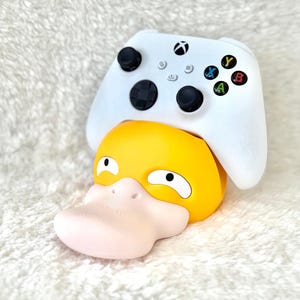 Op de afbeelding: Een witte draadloze videogamecontroller rustend op een gele en lichtroze standaard in de vorm van een personage. De standaard heeft een gestileerde eendachtige kop met een brede snavel, witte ogen met zwarte pupillen en een ronde basis.