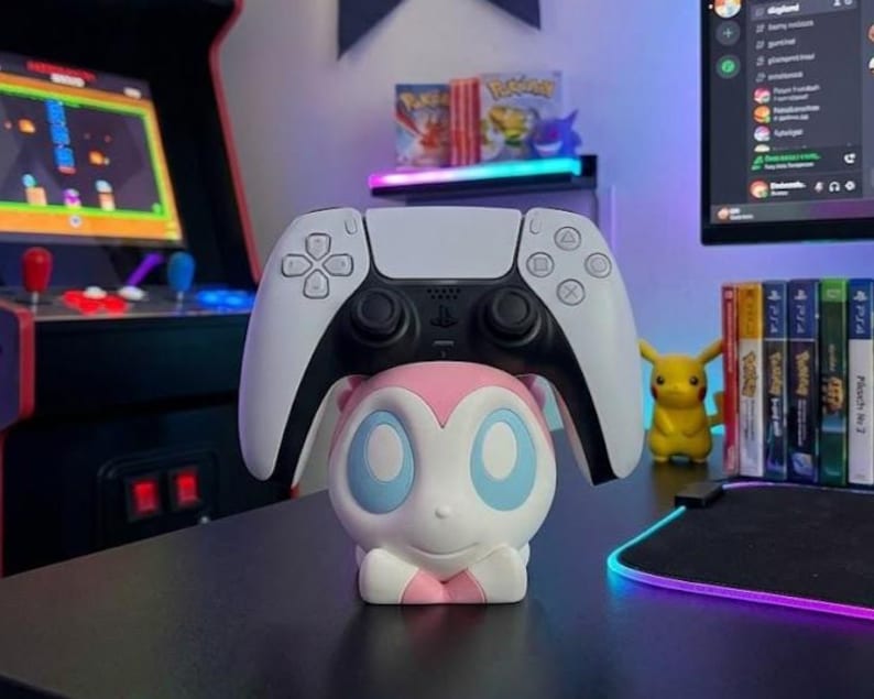 Puede incluir: Un mando blanco y negro de PlayStation 5 descansa sobre una figura de Sylveon rosa y blanco. El mando est&aacute; centrado sobre la figura, con un fondo negro. Una figura de Pikachu y varias carcasas de videojuegos son visibles en el fondo.