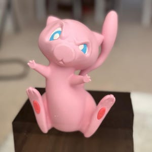 May include: A pink Mew figurine with large blue eyes and a long, slender tail. The character is depicted in a seated position with its arms raised and small red pads visible on the soles of its feet. The surface has a smooth, matte finish.