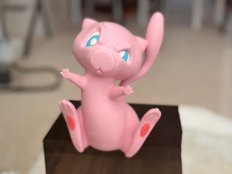 May include: A pink Mew figurine with large blue eyes and a long, slender tail. The character is depicted in a seated position with its arms raised and small red pads visible on the soles of its feet. The surface has a smooth, matte finish.