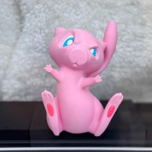 Puede incluir: Una figura rosa de Mew con grandes ojos azules, orejas puntiagudas y una cola larga. El personaje est&aacute; representado en posici&oacute;n sentada con los brazos levantados y pies peque&ntilde;os que presentan almohadillas rosas ovaladas en las plantas.