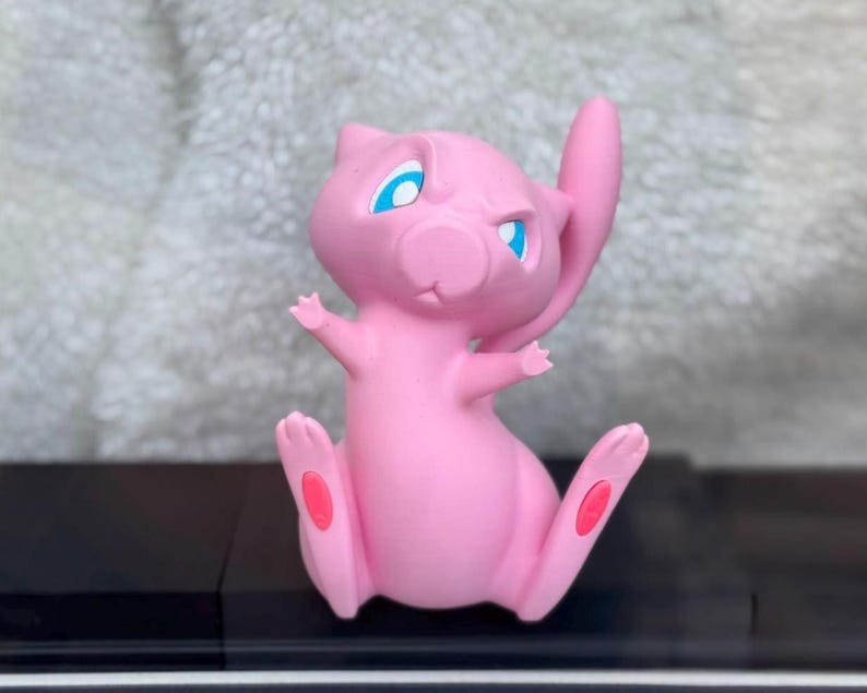 Puede incluir: Una figura rosa de Mew con grandes ojos azules, orejas puntiagudas y una cola larga. El personaje est&aacute; representado en posici&oacute;n sentada con los brazos levantados y pies peque&ntilde;os que presentan almohadillas rosas ovaladas en las plantas.