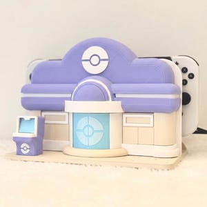 May include: A lavender and cream-colored gaming console dock shaped like a building with a central blue door and a Pok&eacute; Ball emblem. The dock holds a white handheld gaming console with black buttons, and a small matching lavender arcade machine sits to the left.