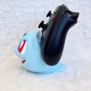 May include: A light blue Bulbasaur-shaped controller stand holding a black gaming controller. The stand features the character's signature red eyes, white teeth, and green bulb detail on the back, providing a stable base for the controller.
