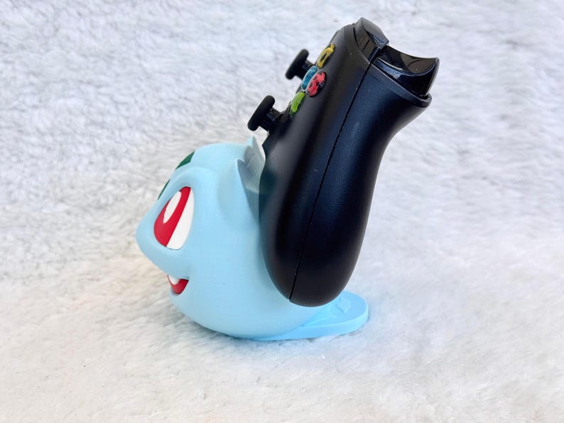 May include: A light blue Bulbasaur-shaped controller stand holding a black gaming controller. The stand features the character's signature red eyes, white teeth, and green bulb detail on the back, providing a stable base for the controller.