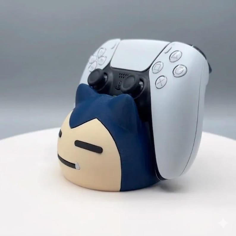 May include: A white PlayStation 5 controller sits on a blue and tan Snorlax-themed controller holder. The holder features a cartoon design with a smiling face. The controller is white with black buttons.